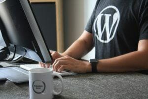 a man wearing a grey wordpress shirt works at a desktop computer with a cup of coffee