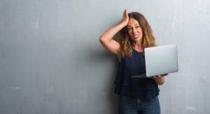 Middle age hispanic woman standing over grey grunge wall using laptop stressed with hand on head, shocked with shame and surprise face, angry and frustrated.