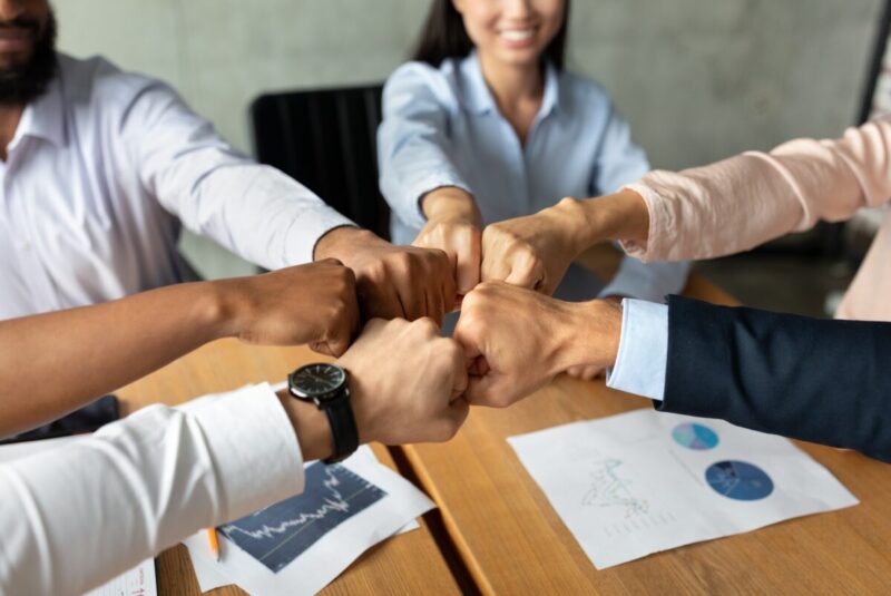 Teamwork Concept. Multiethnic group of business people making fist bumps together while sitting at desk in modern office, company employees joining hands as sign of unity and success, closeup